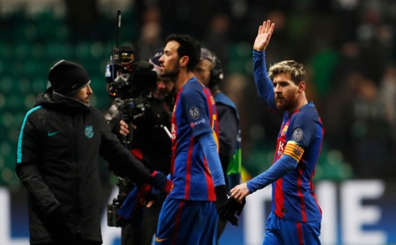 Two men walk across a field as cameras follow them, both dressed in the Barcelona soccer jersey: blue and red stripes. One raises his hand to wave.