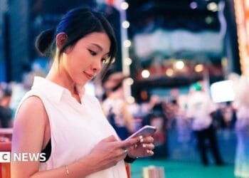 A woman in a white sleeveless top stands in New York City's Times Square at dusk, looking down at the smartphone in her hands