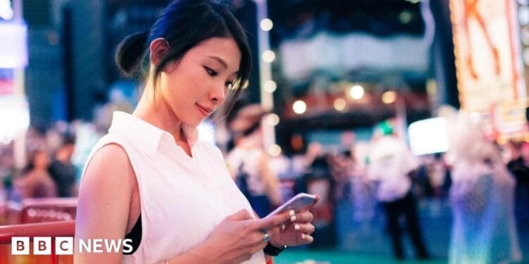 A woman in a white sleeveless top stands in New York City's Times Square at dusk, looking down at the smartphone in her hands
