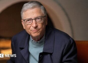 Bill Gates in blue jumper and jacket, sitting by fireplace
