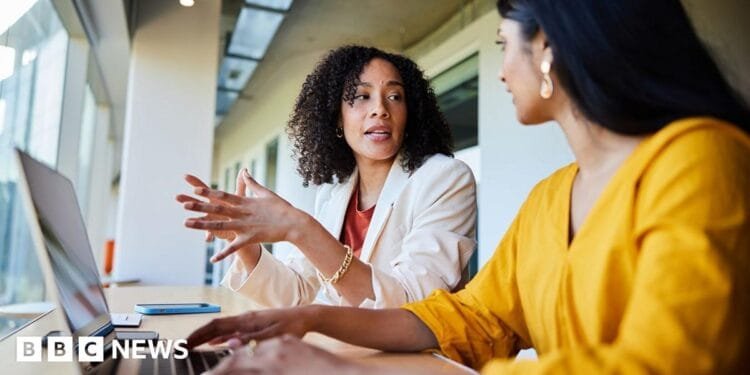 Two women working together in an office, with one gesturing with her hands as she speaks, and the other typing on a keyboard