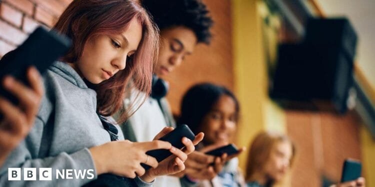 Four school pupils are standing against a brick wall, not talking to each other as they each look down at a phone in their hands